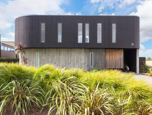 Exterior view of a contemporary two-storey residence with a rounded upper volume clad in dark vertical ribbed metal and a lower level featuring textured pale concrete or timber-like cladding. Narrow vertical windows punctuate the upper facade, while layered native-style grasses and planting occupy the foreground.