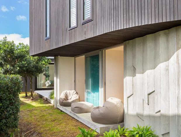 Exterior view of a contemporary two-storey home with a projecting upper volume clad in vertical dark timber boards. Narrow vertical windows punctuate the facade, while a recessed ground-level outdoor sitting area sits beneath the overhang beside a textured pale concrete wall. A landscaped garden with stepping stones and dense ornamental planting surrounds the house.
