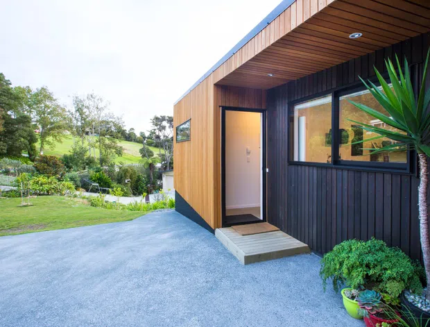 Exterior view of a compact contemporary building with warm timber cladding, dark vertical wall cladding, and a projecting timber-lined roof canopy. A glazed entry door and window open onto a small concrete step, with a gravel forecourt and landscaped garden surrounding the structure.
