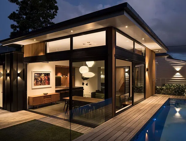 Twilight exterior view of a contemporary, flat-roofed residential extension or pavilion with extensive floor-to-ceiling glazing, dark structural framing, warm timber cladding, and an illuminated open-plan living and dining area visible inside. A timber deck wraps around the building and borders a narrow swimming pool, with integrated exterior lighting and landscaped screening.