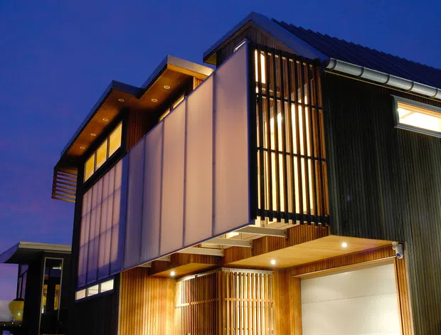 A dusk exterior photograph of a contemporary multi-level residence with a strongly articulated geometric form. The facade combines dark vertical cladding, warm timber soffits and screening, translucent panelled upper walls, extensive glazing, and a large white sectional garage door. Vertical timber and dark metal fins provide privacy and texture, while integrated lighting highlights the entry and recessed outdoor areas. The building is viewed from a paved driveway or forecourt against a deep blue evening sky.