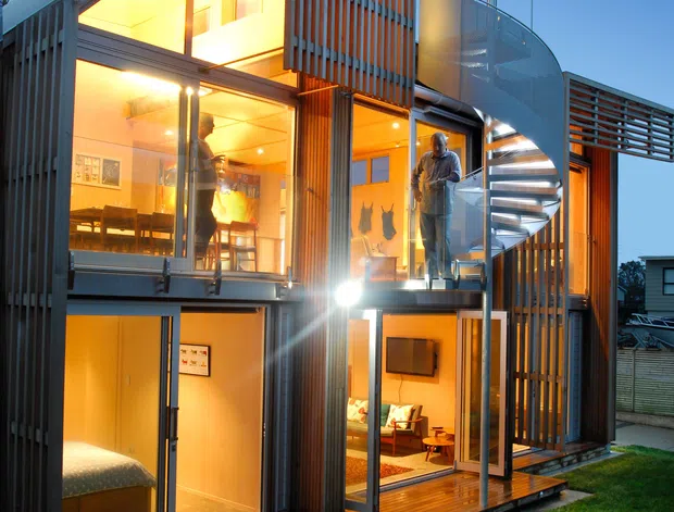 A multi-storey contemporary residential building photographed at dusk, with extensive floor-to-ceiling glazing, warm illuminated interiors, timber decking, vertical timber screening, and a prominent external spiral staircase with curved translucent balustrade. Several living areas and a bedroom are visible through the glazing, opening onto a small lawn and deck.
