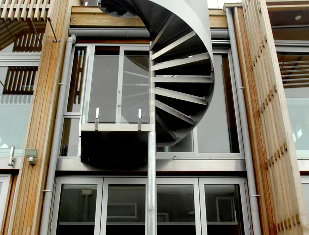 Low-angle exterior view of a contemporary multi-storey building featuring a prominent curved spiral staircase with a perforated white metal balustrade and dark treads. The facade combines extensive aluminium-framed glazing with warm timber cladding, vertical timber screening, balconies, exposed services, and a central structural pole.