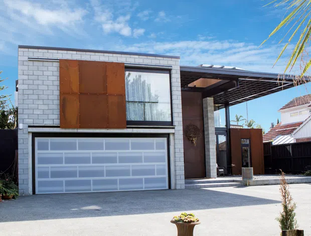 Exterior view of a contemporary two-storey residence with a white masonry-block facade, large translucent sectional garage door, rusted steel feature panel, and a deep covered entry or carport canopy. The home has strong geometric forms, dark framing, and a paved driveway, with neighbouring residential buildings visible at the sides.