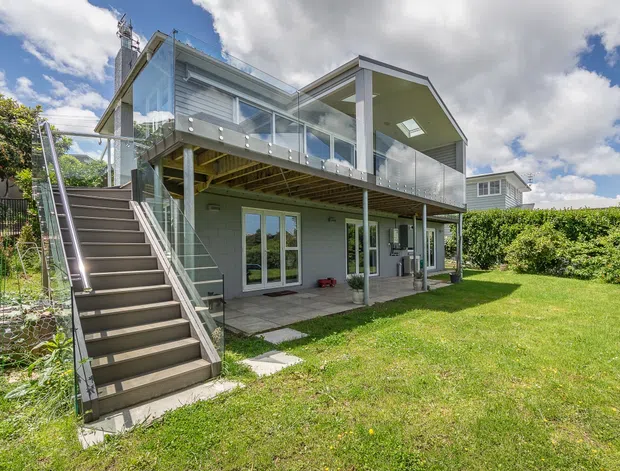 Exterior rear view of a contemporary multi-level home with grey horizontal cladding, broad glazed balcony balustrades, an elevated upper living level, and a covered ground-floor outdoor area. A wide external stair connects the lawn to the upper deck, with manicured hedges and a bright partly cloudy setting.