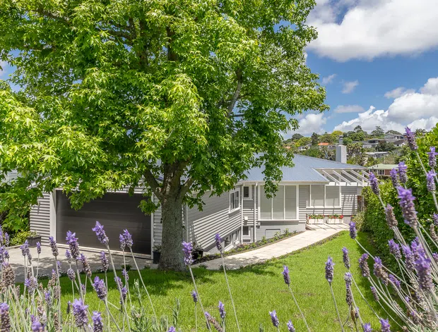 Exterior view of a low-rise suburban home with light grey horizontal cladding, a shallow pitched metal roof, attached garage, and covered entry walkway. Mature trees and purple flowering plants frame the landscaped front yard, with neighbouring homes visible on the sloping site behind.
