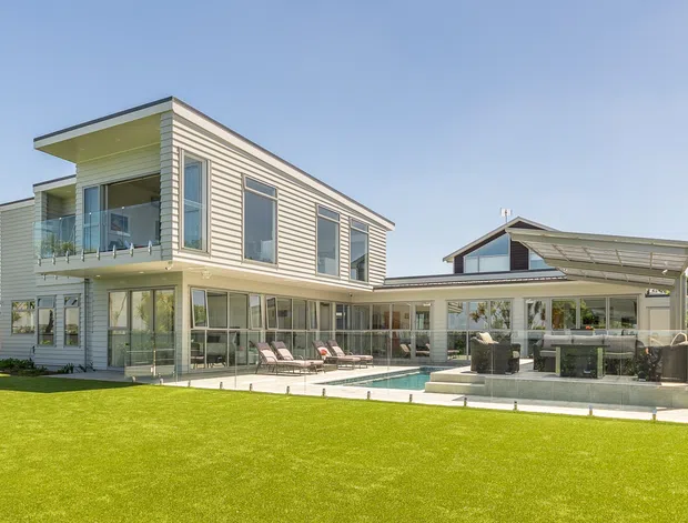 A bright exterior view of a contemporary multi-level home with light horizontal weatherboard cladding, a low-pitched roof, extensive glazing, and a covered outdoor entertaining area. The house opens onto a broad manicured lawn with a small swimming pool, outdoor seating, and a sheltered kitchen or barbecue zone.