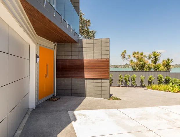 Contemporary residential exterior entry and driveway, featuring a bright orange front door, large light-panel wall cladding, grey concrete blockwork, horizontal timber screening, a timber-lined soffit, and upper-level glass balustrades. A landscaped boundary and turquoise water are visible beyond the property.