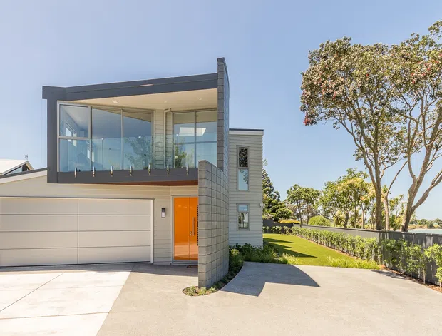 Contemporary two-storey residential facade with a wide integrated garage, large upper-level glazed room or balcony enclosure, grey rendered walls, a curved glass-block feature wall, and a vivid orange entry door. The home sits beside a paved driveway and landscaped boundary with mature trees.