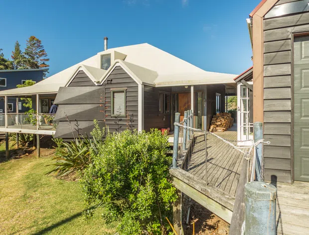 Exterior view of a compact multi-building residential property with dark grey horizontal weatherboard cladding, white pitched roof forms, raised timber decking and a fenced entry walkway. The composition includes a prominent garage or outbuilding at right, landscaped garden planting, and neighbouring cottage-like structures under clear daylight.