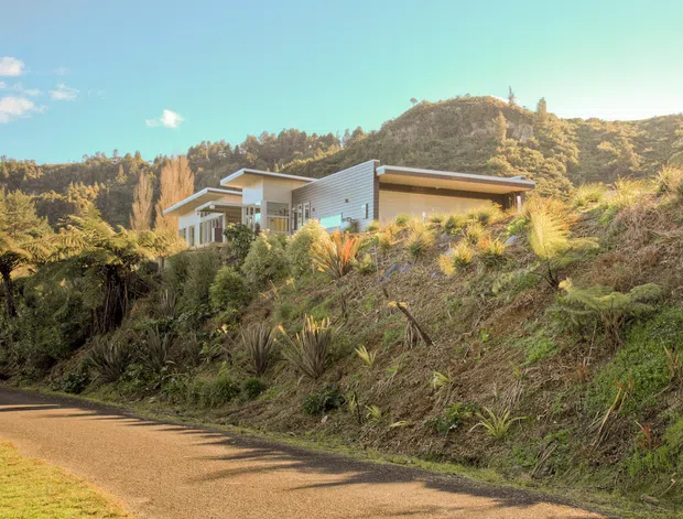 Exterior view of a contemporary single-storey home set into a densely planted, sloping landscape. The house features pale horizontal cladding, broad roof overhangs, extensive glazing and a low-profile form, with a paved driveway or walkway in the foreground and wooded hills behind.