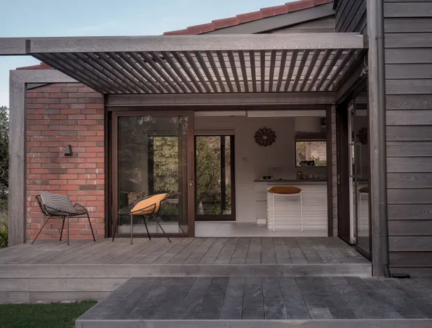 A contemporary single-storey residential extension viewed from the garden, featuring a dark timber-clad facade, red brick walls, a timber deck, and a pergola with closely spaced overhead slats. Large glazed doors open to a compact interior bar or kitchen area, while outdoor seating sits beneath the covered terrace.