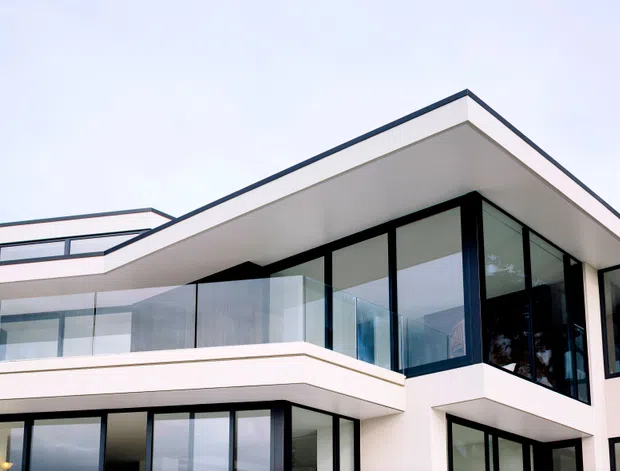 Exterior view of a contemporary multi-level residence with crisp white rendered walls, strong horizontal roof overhangs, extensive black-framed glazing, and a glass-balustraded balcony. The flat-roofed form and projecting upper volume create a clean, geometric facade.