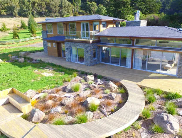A modern two-storey house set within a landscaped rural or bush setting. The home features a low-pitched roof, extensive glazing, timber-framed windows and doors, stone-clad lower-level walls, timber decking, and a curved timber boardwalk winding through a landscaped garden of rocks and grasses.