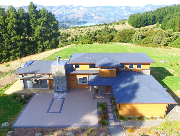 Aerial view of a contemporary single-storey hillside residence with multiple low-pitched roof volumes, warm timber cladding, grey metal roofing, a prominent stone chimney, broad paved outdoor areas, and an attached garage. The home is surrounded by landscaped grounds and dense vegetation, with a broad mountain and valley landscape visible beyond.