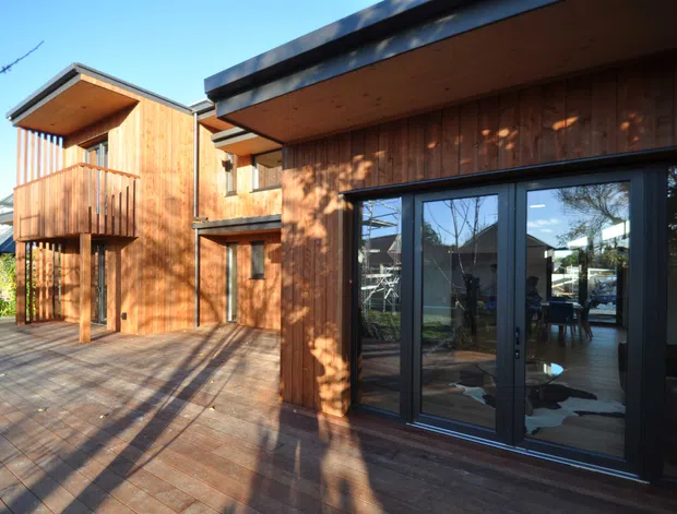 Exterior view of a contemporary multi-level home with extensive warm timber cladding and decking. The building has a projecting upper balcony, deep roof overhangs, dark-framed glazed folding or sliding doors, and a sheltered outdoor deck. Interior dining and living furnishings are visible through the glazing.