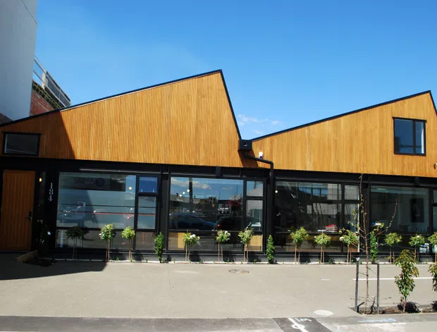 Exterior view of a contemporary low-rise building with two asymmetrical pitched roof volumes, warm vertical timber cladding, dark window and door frames, and a glazed ground-level frontage opening onto a paved forecourt with young planted trees. Urban buildings are visible around the site.