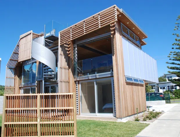 Exterior view of a compact multi-level contemporary building with prominent timber batten screening, a curved external stair, broad glazing, balconies, and a projecting upper roof. The structure sits beside a paved walkway with landscaped residential surroundings under a clear blue sky.