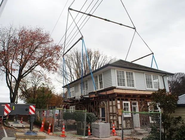 A traditional two-storey timber-clad house is being lifted or repositioned by a large overhead rig at an active construction site. The upper level has white weatherboard cladding, multi-pane windows, and a dark hipped roof, while the lower level is exposed with visible structural framing. Temporary fencing, concrete blocks, traffic cones, and lifting equipment surround the building. Overcast outdoor conditions and mature trees form the background.