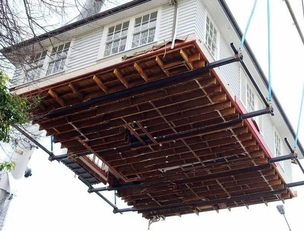 Low-angle exterior view of a raised timber-framed deck or balcony structure being supported by temporary steel scaffolding or bracing. The upper storey has white horizontal weatherboard cladding, white-framed multi-pane windows, and dark trim, while the underside exposes timber joists and framing.