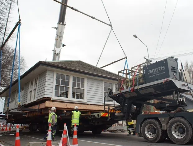 A traditional timber-clad house being lifted and transported by a heavy vehicle on a city street. The house has horizontal weatherboard siding, a hipped shingle roof, and prominent multi-pane windows. Construction workers, lifting equipment, traffic cones, and utility wires surround the move.
