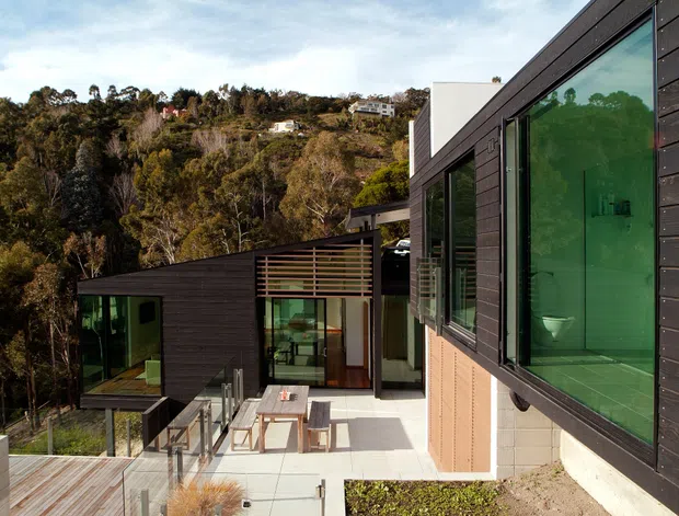 Exterior view of a contemporary multi-level hillside home with dark horizontal cladding, extensive glazing, and connected outdoor terraces. The house is embedded into a wooded, sloping site, with a central glazed entry or living area and a large projecting window revealing a bathroom fixture inside. Timber decking, paved terraces, outdoor furniture, and landscaped planting are visible.