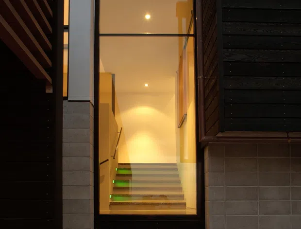 A narrow, modern entry passage viewed from outside, framed by dark horizontal timber cladding and pale masonry walls. A full-height glazed entry door reveals a warmly lit interior stairway with floating or open-riser steps, glass or slim metal balustrade, and recessed lighting. The composition emphasizes vertical glazing, contrasting dark and light materials, and a compact urban architectural entrance.