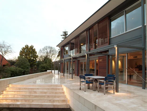 Exterior view of a contemporary multi-level residence with a broad paved terrace, illuminated stone steps, floor-to-ceiling glazing, dark metal framing, timber louvred screens, and pale masonry cladding. Outdoor dining furniture sits on the terrace, with landscaped suburban surroundings visible beyond.