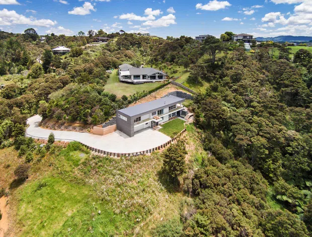 Aerial view of a contemporary multi-level residential house set on a heavily vegetated, elevated and sloping site. The home has a dark grey and white exterior, a broad low-pitched roof, extensive glazing, surrounding concrete terraces and a winding driveway. Other homes, rolling greenery and distant hills are visible in the background.