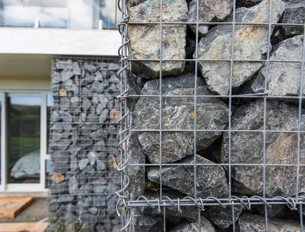 Close-up architectural detail of gabion walls filled with irregular grey and brown natural stones held in galvanized wire mesh cages. A contemporary building facade with large glazed openings and timber decking is visible behind, emphasizing the textured stone construction.