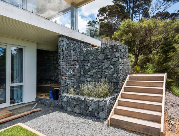 Exterior view of a contemporary multi-level residence set among dense bushland. The facade combines white rendered walls and large glazed doors with substantial gabion stone walls, raised planters, a timber stair, and a glass-balustraded upper terrace.