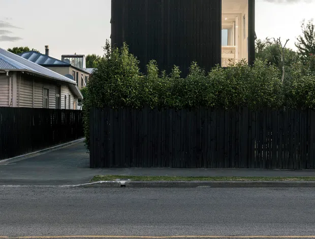 Street-facing view of a compact contemporary two-storey house behind a tall black vertical timber fence. The dark box-like upper volume has black vertical cladding and a large recessed illuminated opening, while neighbouring timber-clad houses and planting frame the scene. The image is photographed from across a residential street in soft overcast evening light.