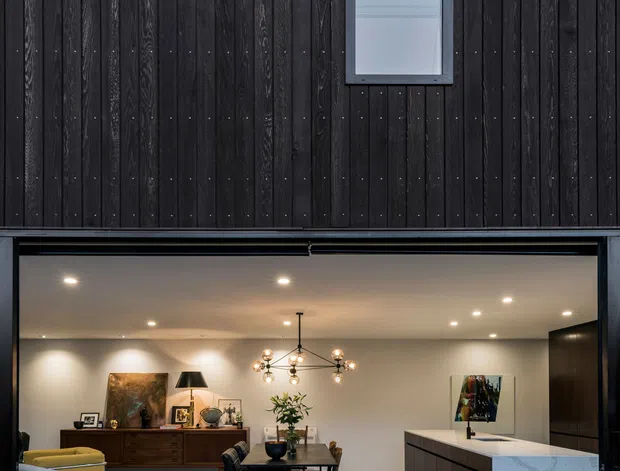 Contemporary residential facade with dark vertical timber cladding and two small square windows above a wide open glazed garage-style opening. The opening reveals a refined open-plan interior combining a dining area, living seating, timber sideboard, decorative lighting, and a modern kitchen island with a white stone benchtop. The composition contrasts black timber exterior surfaces with warm neutral interiors and dark joinery.