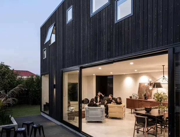Exterior dusk view of a contemporary two-storey home with a dark vertical timber-clad facade, asymmetrical rectangular windows, and expansive ground-floor glazing opening onto a paved outdoor area. The illuminated interior clearly shows an open-plan living and dining space with sofas, dining table, pendant lighting, and built-in timber joinery. The facade and glazing dominate the image.