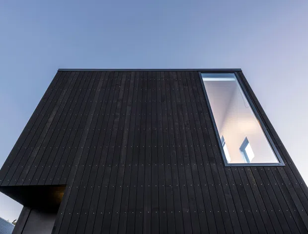 Low-angle exterior view of a sharply composed modern building facade clad in dark vertical panels. A tall, narrow glazed opening or skylight interrupts the facade, revealing a bright interior void and a small window beyond. The image emphasizes the dark cladding, strong geometric lines, and dramatic contrast against a clear blue sky.