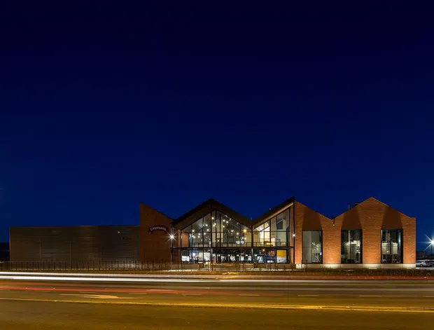 Night-time exterior photograph of a contemporary multi-purpose building viewed across a busy road. The long low facade combines dark horizontal cladding, warm red-brown brick volumes, large glazed gabled sections, and brightly illuminated interiors. Strong street lighting and vehicle light trails emphasize the urban roadside setting.