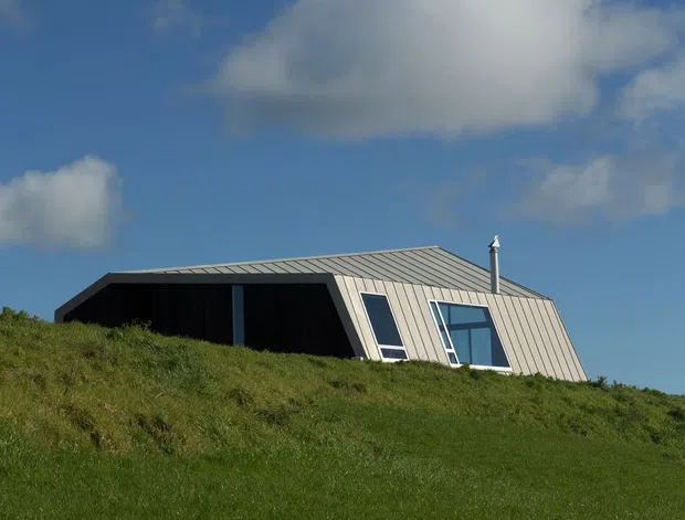 Exterior photograph of a low-profile modern house partially embedded in a grassy sloping site. The building has a sharply angled, folded roof form with grey metal roofing/cladding, dark recessed wall surfaces, large slanted windows, and a visible metal chimney. The image is dominated by the green hillside and blue sky with clouds.