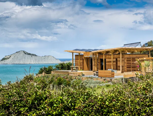 A low-slung timber-clad coastal dwelling set beside vivid turquoise water, with extensive timber decking, pergola-like roof framing, solar panels, and louvred timber screens. A distinctive pale rocky island or headland is visible offshore, while dense coastal planting fills the foreground. The architecture has a strongly site-responsive, relaxed and sustainable coastal character.