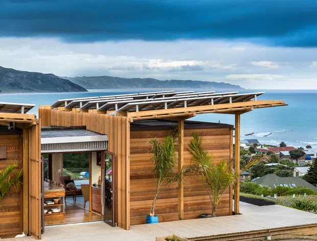 Exterior view of a contemporary timber-clad coastal residence with a broad, layered low-pitched roof and extensive outdoor deck. The home overlooks a bay or ocean, with rugged coastal hills and neighbouring houses visible beyond. Large glazed openings connect the deck to the interior, while palm plantings and warm timber surfaces reinforce the relaxed seaside setting.