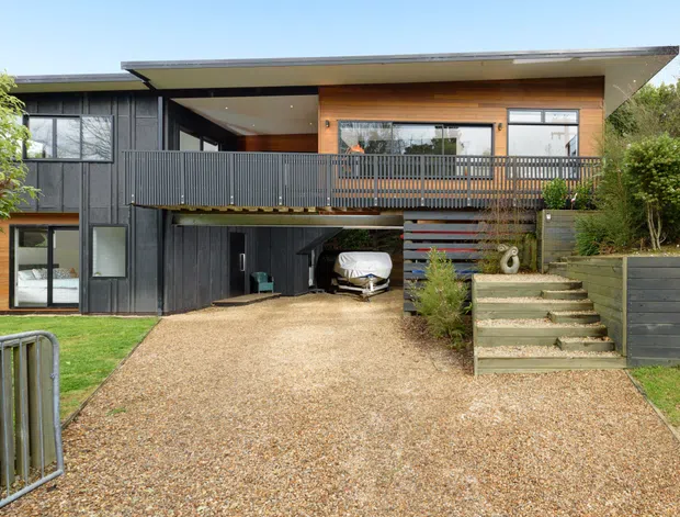 Exterior view of a contemporary elevated home with dark vertical cladding, warm timber wall lining, broad low-pitched roof, upper-level balcony, and a partially enclosed undercroft carport. A gravel driveway leads to the house, with landscaped retaining walls and steps.