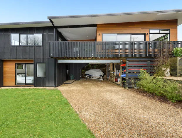 Exterior view of a contemporary split-level home with dark vertical cladding, warm timber wall panels, broad roof overhangs, and a raised upper-level balcony. The lower level includes glazing and an open carport or storage area, with a gravel driveway, lawn, retaining walls, and steps visible.