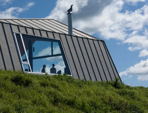 Exterior view of a modern building integrated into a steep grassy hillside. The visible facade has dark standing-seam metal cladding, a sloping roof, a large angular glazed window, and a metal chimney, set against a blue sky with clouds.