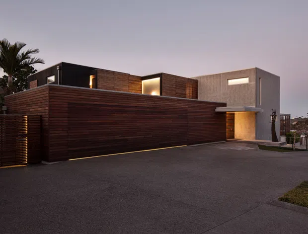Twilight exterior view of a sophisticated contemporary residence with strong horizontal composition. Dark timber-clad walls and gates form the foreground, with contrasting smooth grey rendered volumes, recessed warm lighting, narrow horizontal windows, and a flat-roofed, cubic massing. A broad paved driveway dominates the approach, with limited landscaping and palm vegetation at the edges.