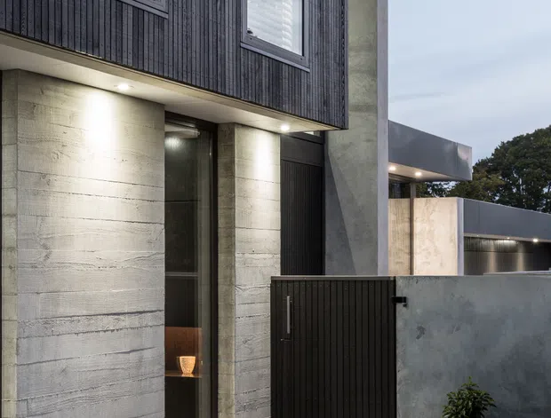 Contemporary multi-level residential facade viewed from the front walkway, featuring dark vertical timber cladding, board-formed concrete walls and columns, slim dark window frames, projecting box-like upper volumes, and a black vertical-slat pedestrian gate. A pale tiled path leads through sparsely planted garden beds toward the recessed entry, with warm architectural lighting beneath the overhangs.