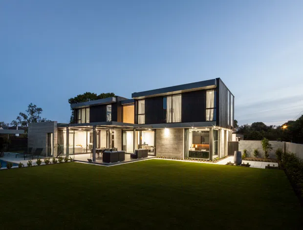 Dusk exterior view of a contemporary multi-storey residence with a strong rectilinear form, flat roof sections, extensive floor-to-ceiling glazing, dark vertical upper-level cladding, and exposed concrete lower-level walls. A covered outdoor terrace opens onto a broad lawn, with a swimming pool visible to the left and boundary fencing surrounding the property.
