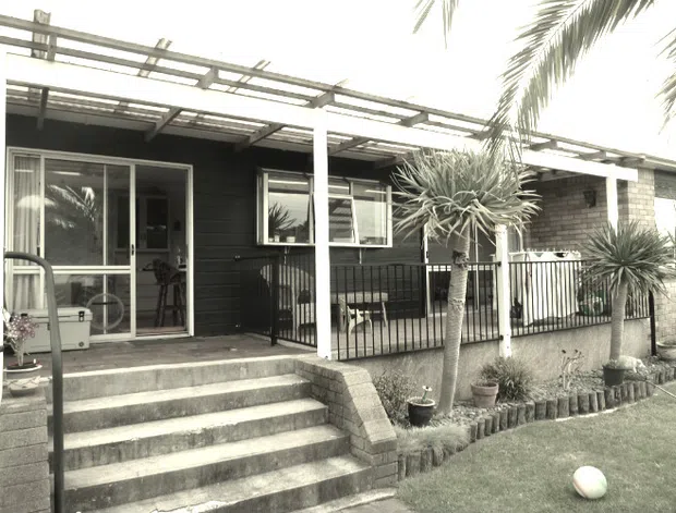 Single-storey residential exterior with dark horizontal cladding, a broad covered terrace or entry veranda, white posts and railings, large glazed doors and windows, and a stepped concrete approach. The setting includes a small landscaped garden with palms and sculptural plants. The image has a muted, slightly monochrome architectural-photography appearance.