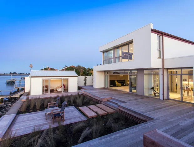 Contemporary multi-level waterfront residence with crisp white rendered walls, expansive glazing, and red-brown roof trim. Broad timber decking forms terraces around landscaped garden beds, with a small detached pavilion by the water and a private dock visible at the left.