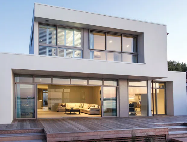 Exterior view of a two-storey contemporary residence with a clean white rendered facade, flat roof, broad floor-to-ceiling glazing, timber-framed windows and doors, and a large timber deck. The open lower level reveals a warm living area with a sectional sofa and dining space, while the upper level contains bedrooms or private rooms.