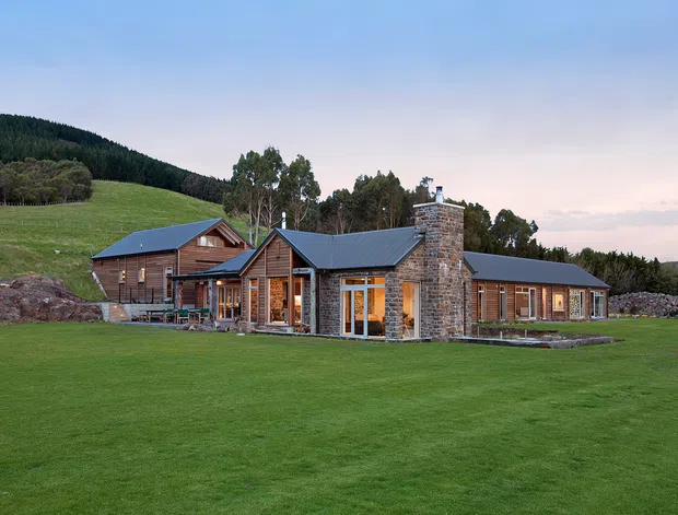 A sprawling single-storey rural house set in a broad manicured lawn, with multiple timber-clad wings, dark pitched metal roofs, and a prominent rough stone chimney. Large glazed openings reveal warm interior lighting, while wooded hills and open pasture form the surrounding landscape.
