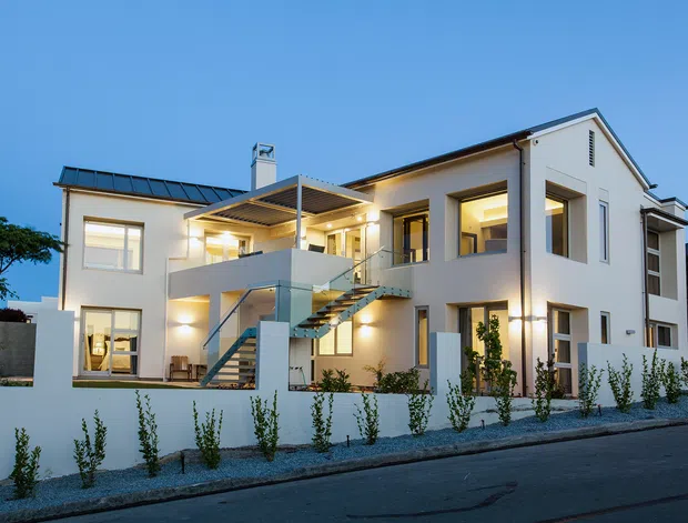 A contemporary two-storey white rendered residence photographed at dusk, featuring a combination of gabled and low-pitched roof forms, large rectangular windows, a projecting upper-level terrace, and an external open-riser stair with glass balustrades. Warm interior and exterior lighting highlights the clean geometric facade, landscaped front boundary, and paved street frontage.